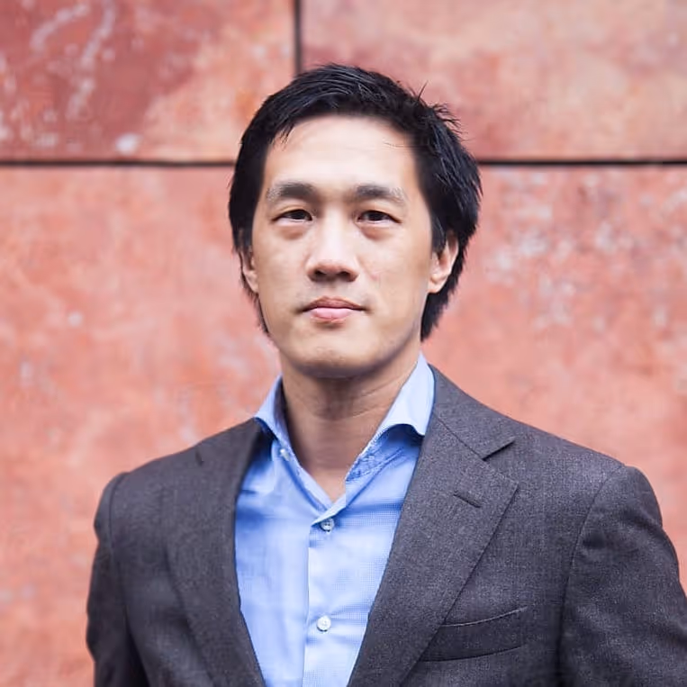 Portrait of a confident man wearing a gray suit jacket and light blue shirt against a reddish stone wall background.