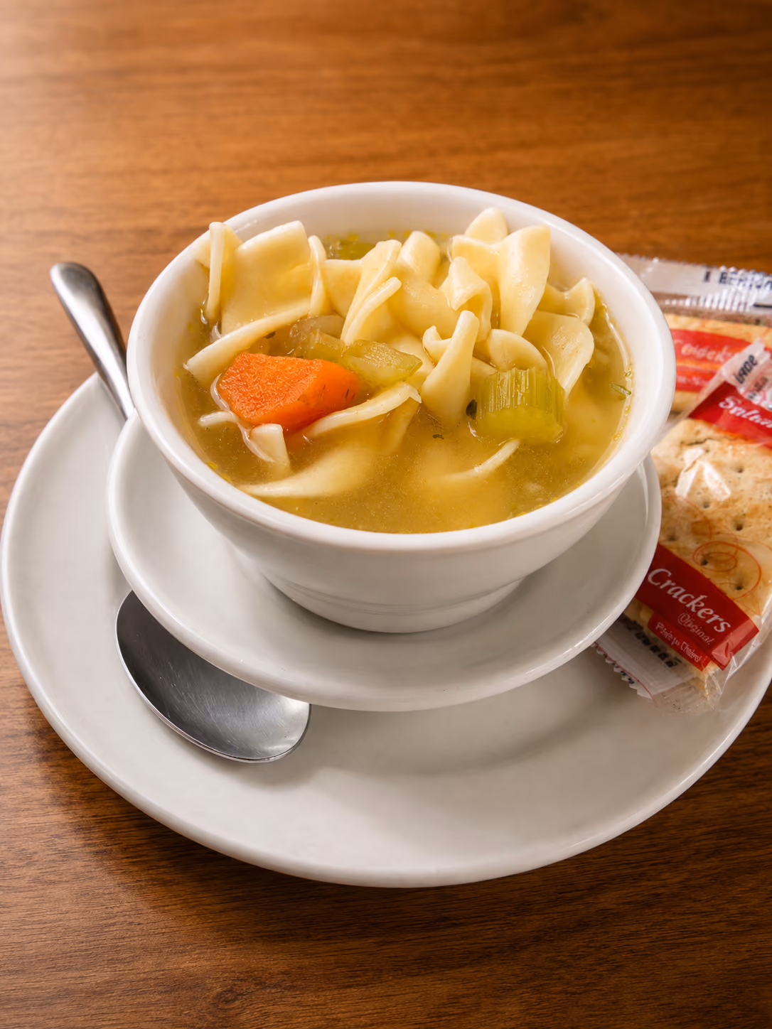 White bowl of chicken noodle soup with carrots and celery, spoon, and cracker packet on a wooden table.