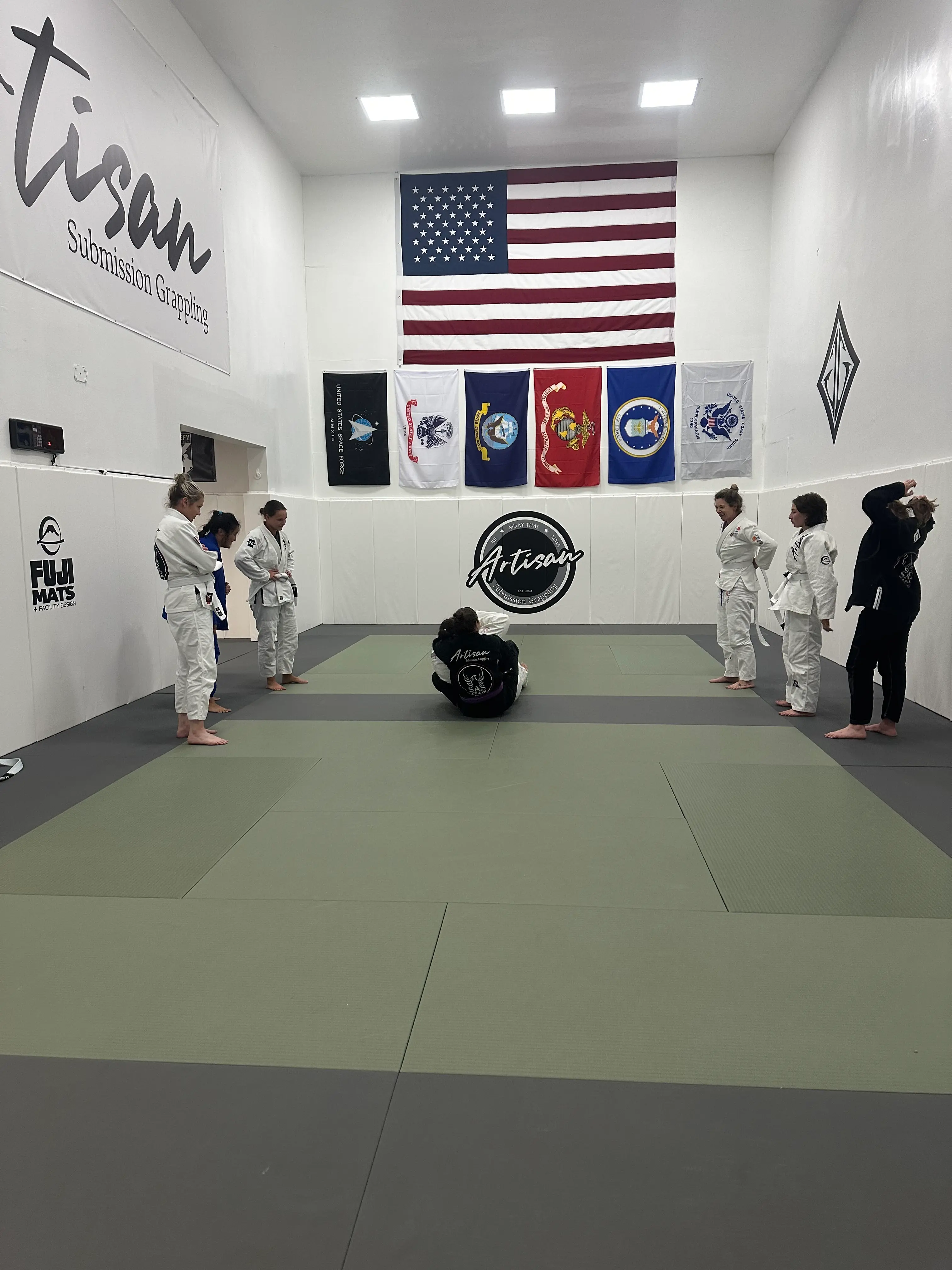 A group of women in martial arts uniforms stand around a training mat, observing two women demonstrating a ground grappling technique during a ladies-only self-defense class at Artisan MMA.