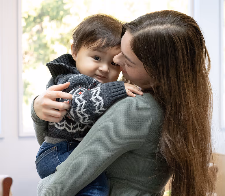 Woman with long brown hair holding and embracing a toddler wearing a patterned sweater inside a brightly lit room.