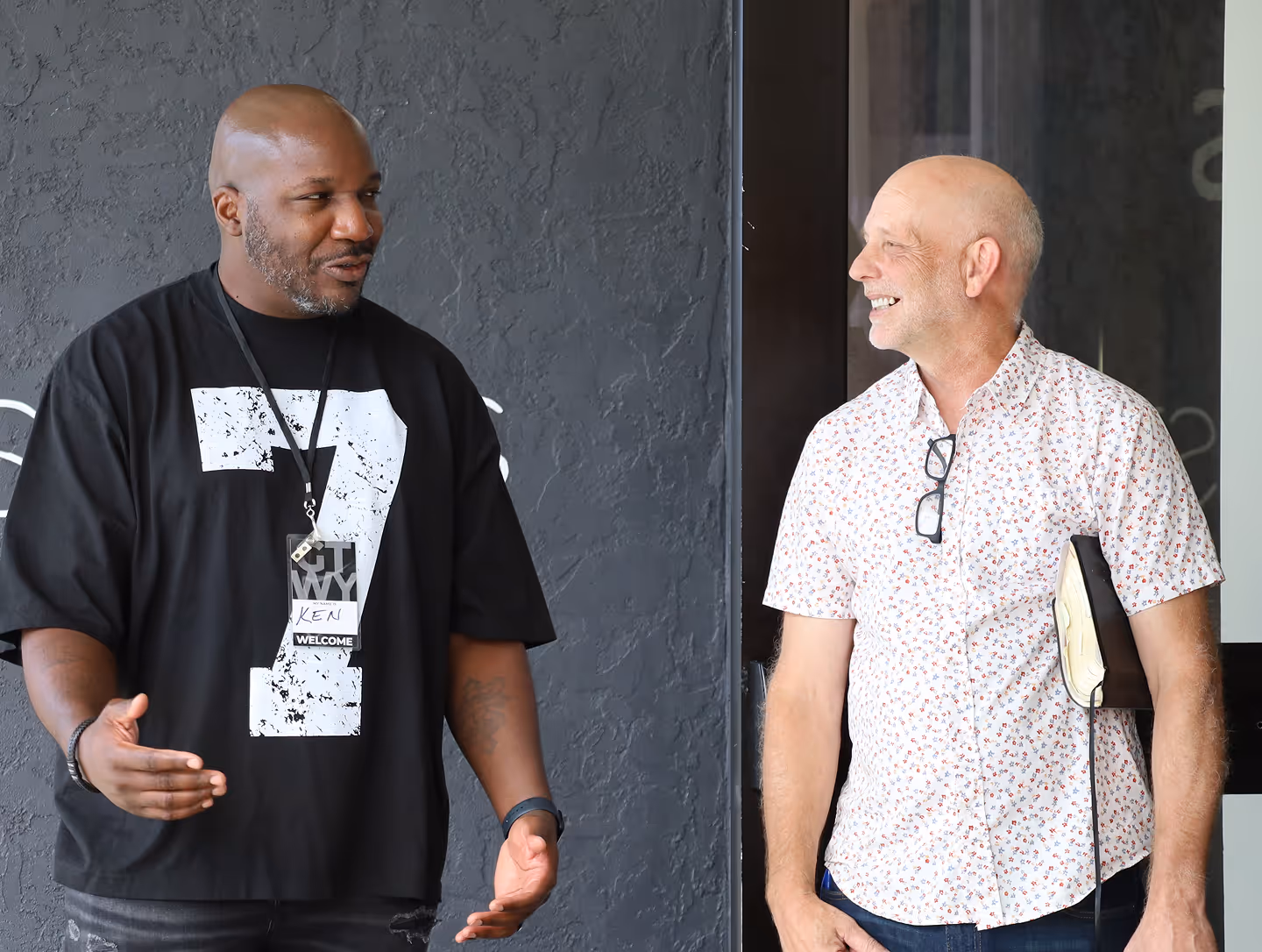 Two men smiling and talking, one wearing a black shirt and name tag, the other holding a book and wearing glasses on his shirt.