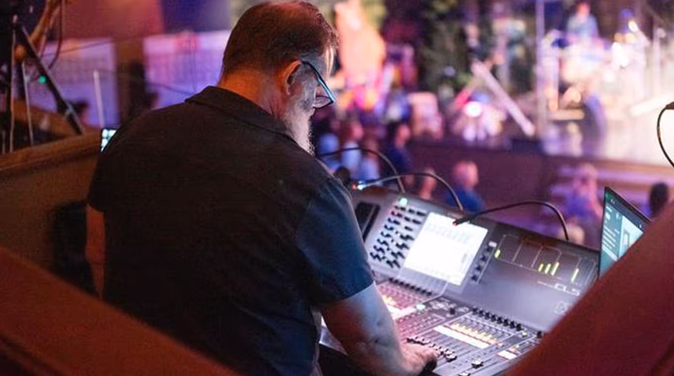 Man with glasses and beard operating a sound mixing console at an event with blurred audience in the background.