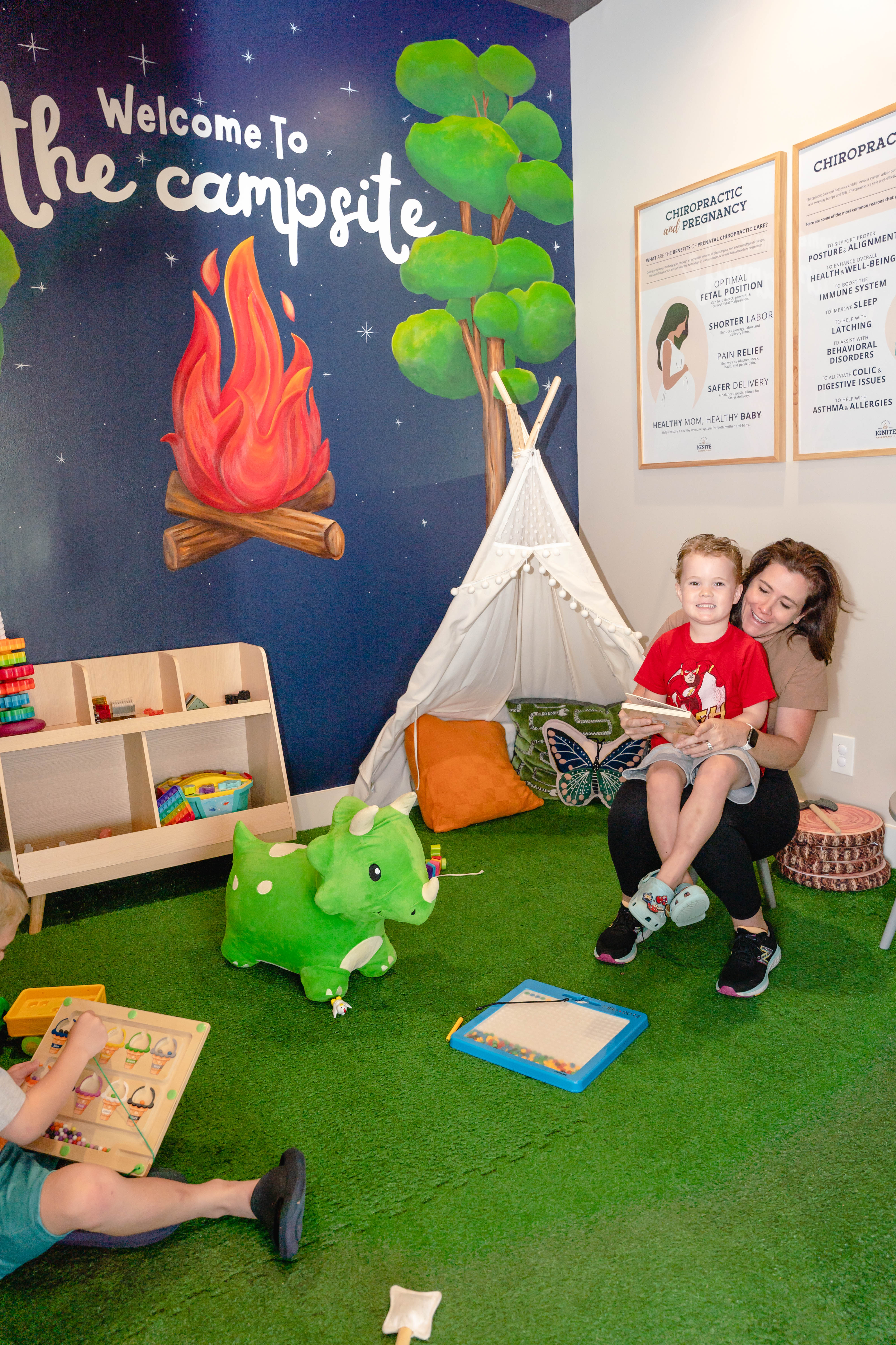 Child-friendly chiropractic waiting area with play tent, toy dinosaur, books, and family enjoying playful learning activit...