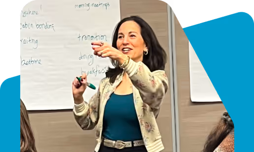 Woman in floral cardigan and blue top standing and pointing while speaking in a meeting room with handwritten notes on paper behind her.
