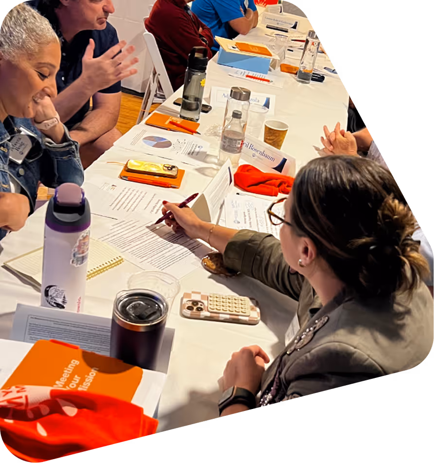 People seated around a table engaged in discussion with papers, notebooks, phones, and water bottles in front of them.