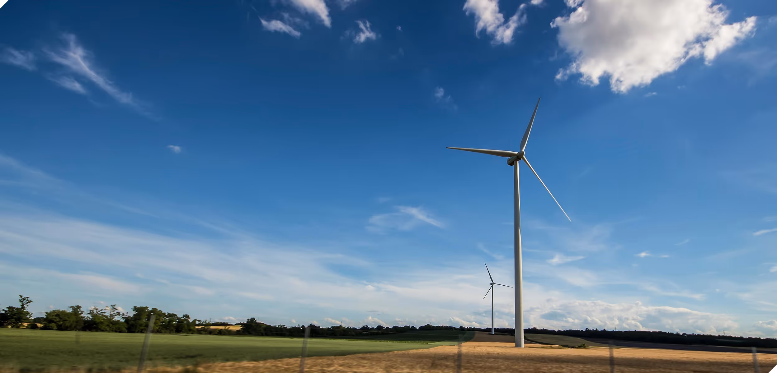 Two large wind turbines in a rural field under a blue sky with scattered clouds.