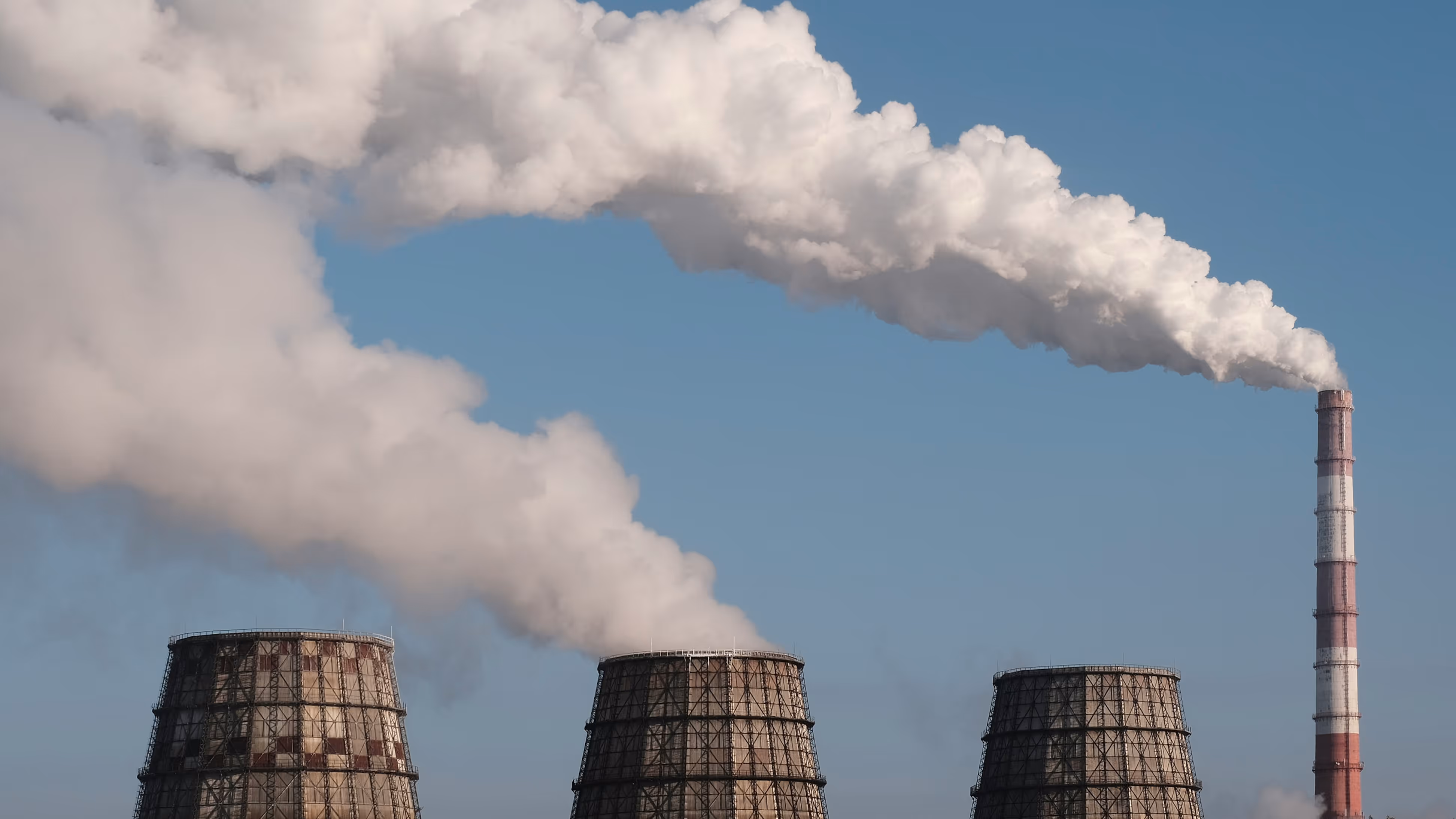 Industrial cooling towers and a tall smokestack emitting thick white smoke against a clear blue sky.