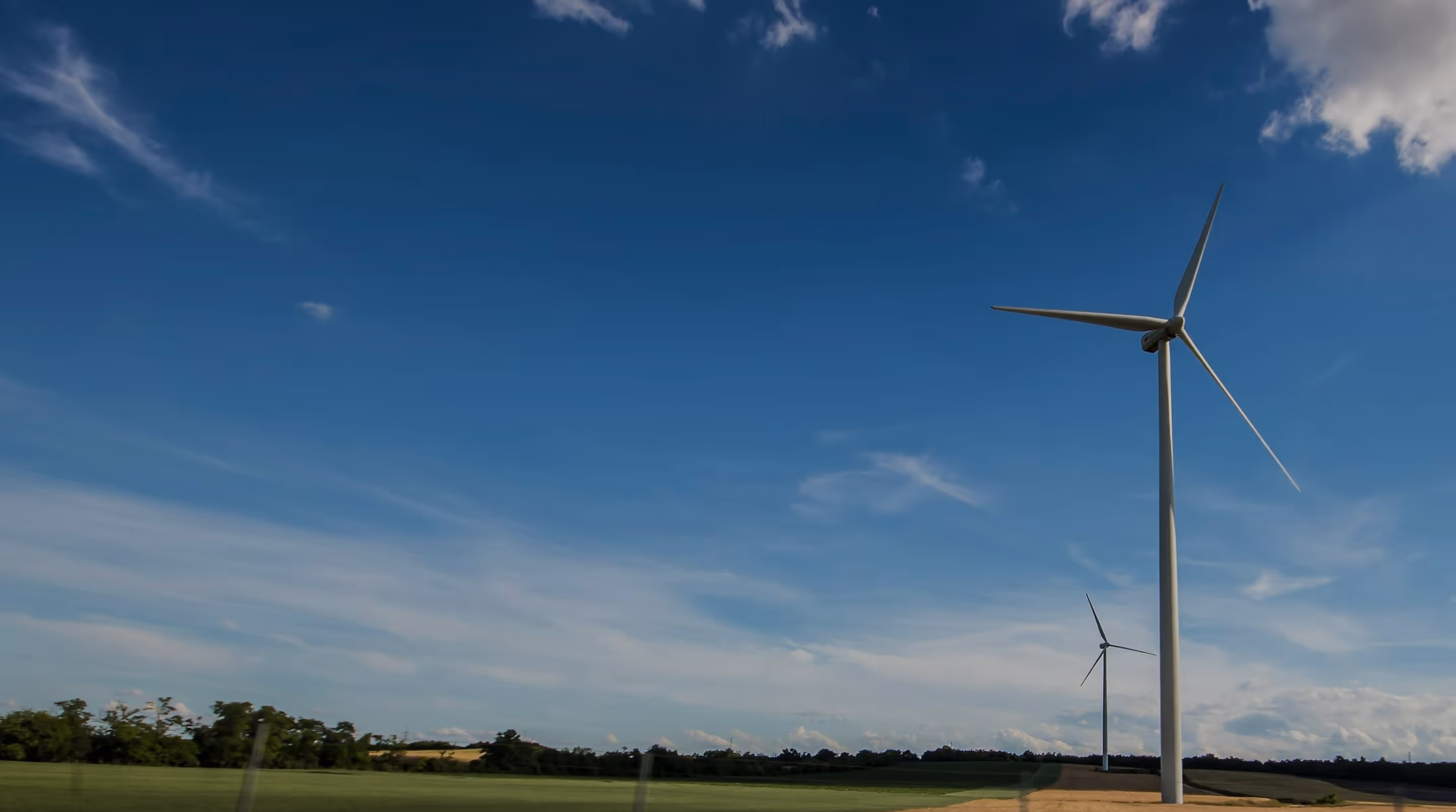 Two large wind turbines in a green field under a partly cloudy blue sky.
