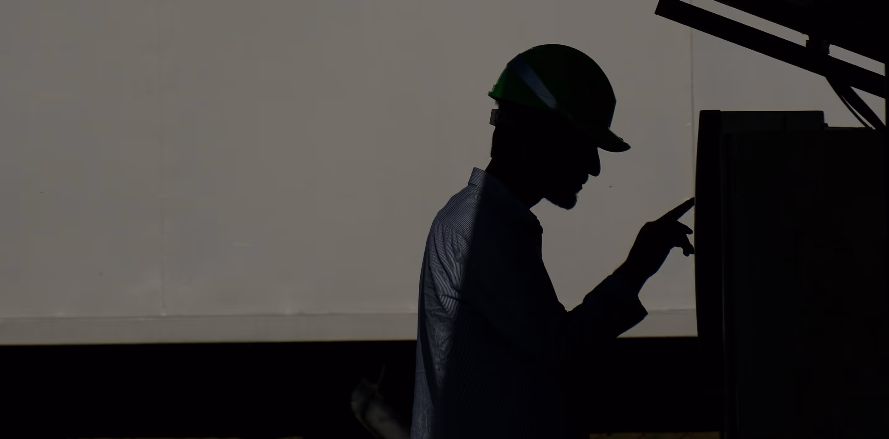 Silhouette of a construction worker wearing a hard hat and pointing at a control panel or machine.