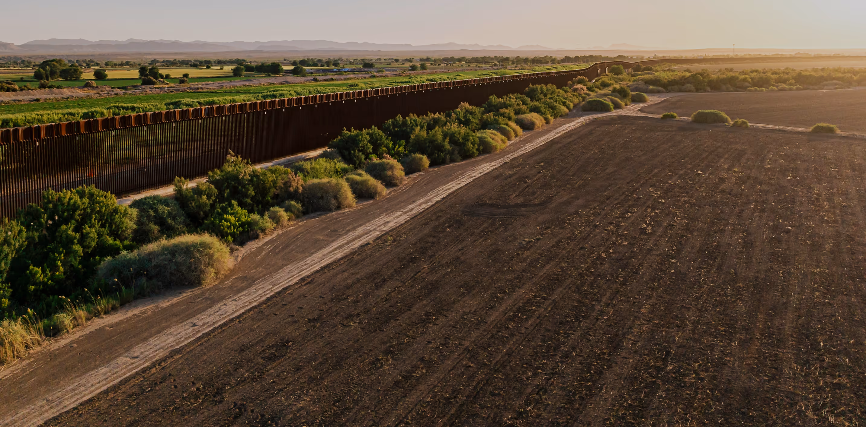 Long border fence running through a rural landscape with greenery on one side and plowed field on the other during sunset.