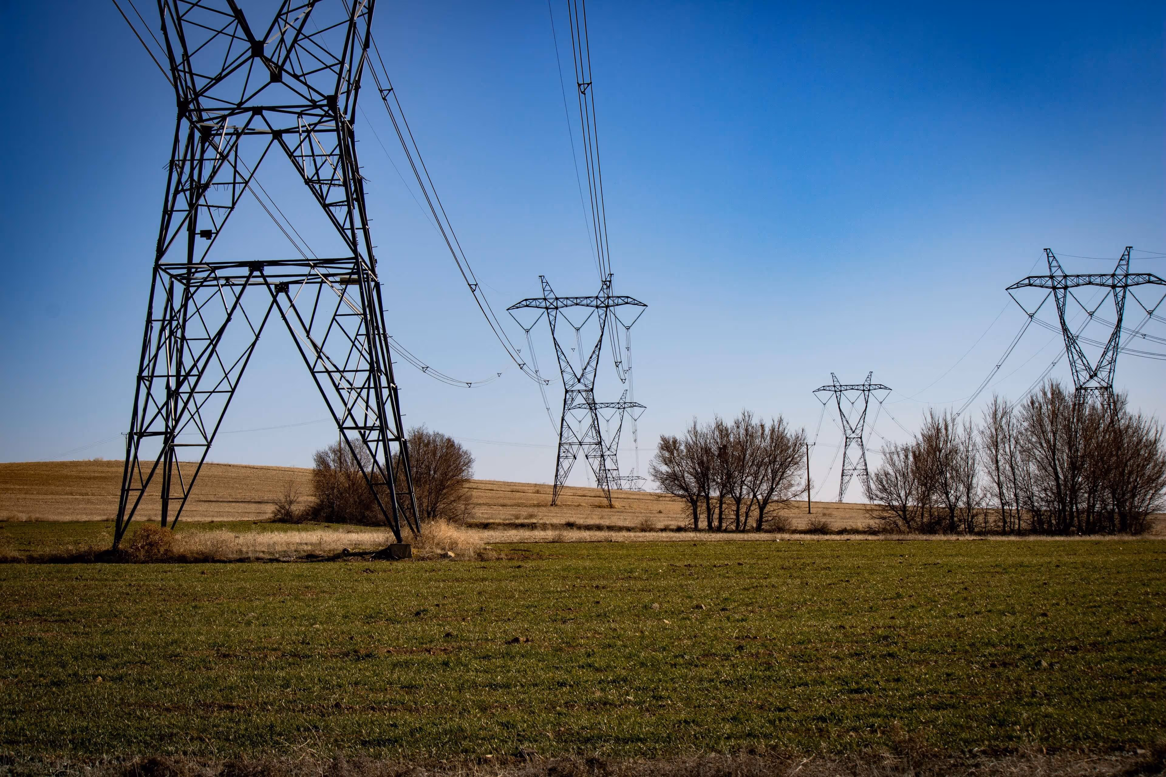 A series of metal electrical transmission towers stretching across a green field and dry hills under a clear blue sky.