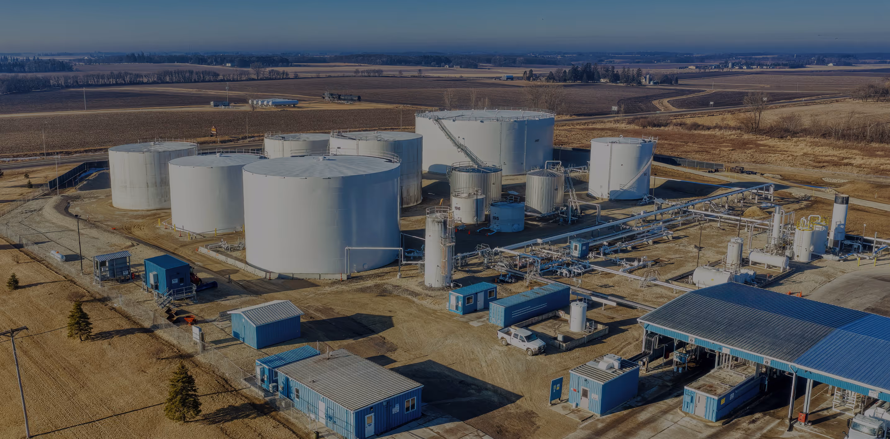 Aerial view of an industrial site with multiple large white storage tanks, blue utility buildings, and pipelines in a rural area.