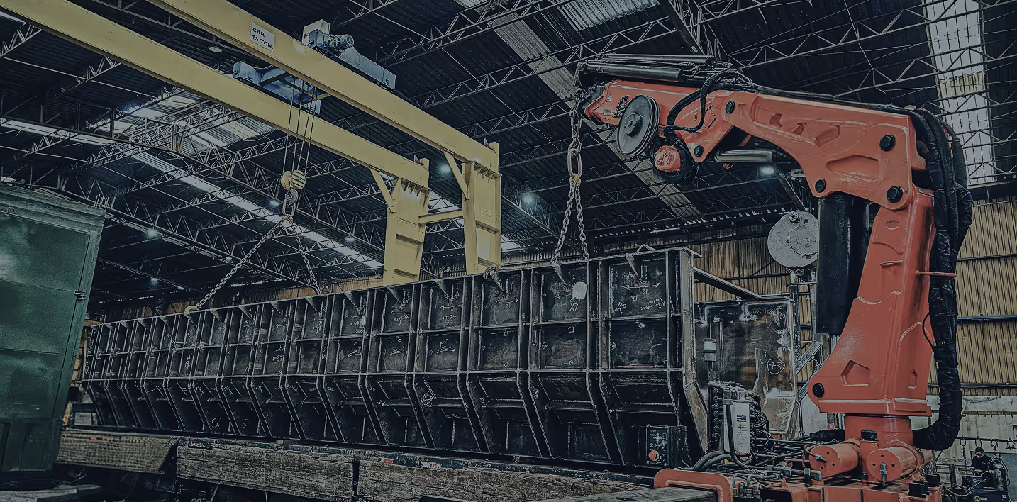 Industrial warehouse interior with a large metal structure being lifted by yellow overhead crane and an orange robotic arm.