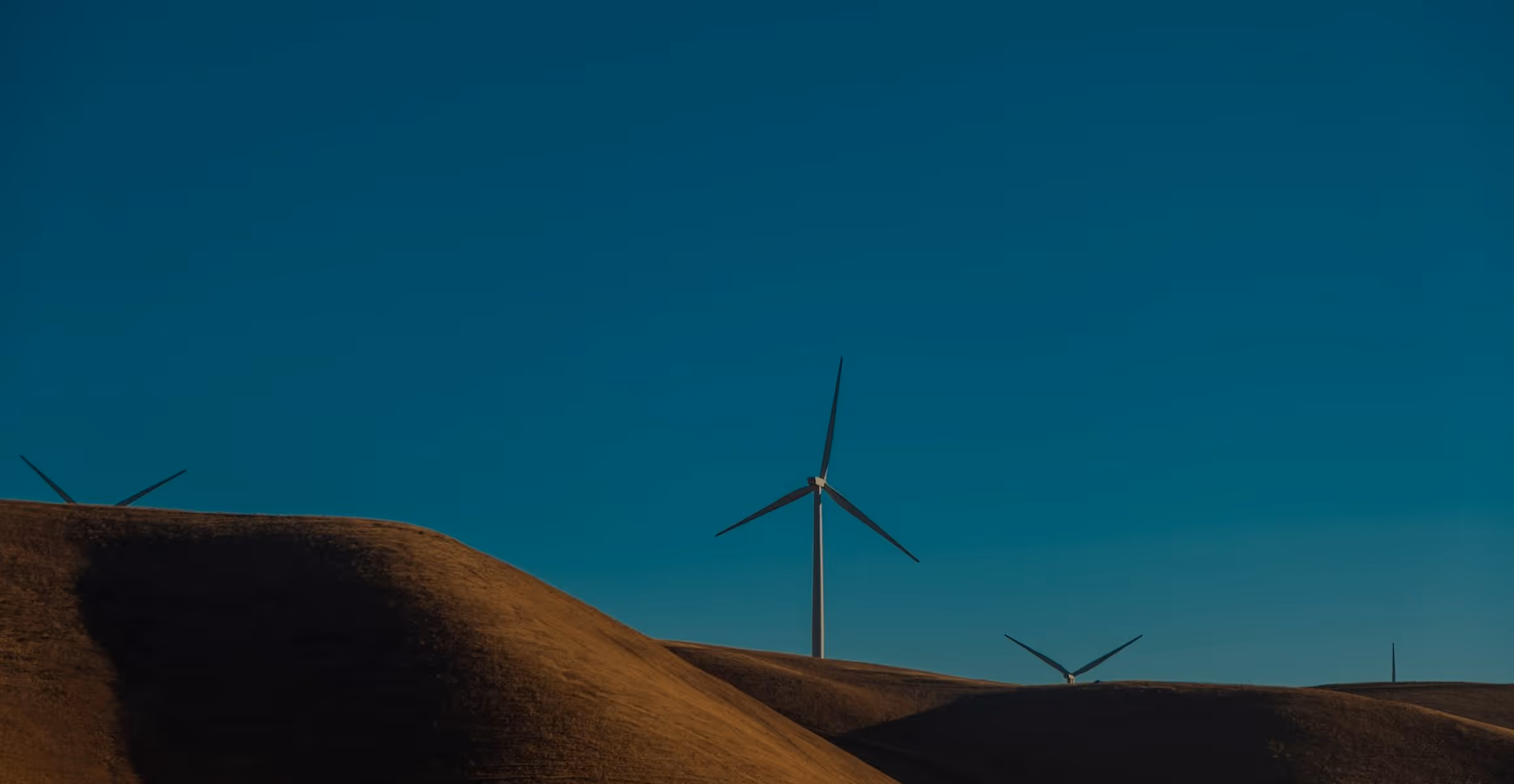 Wind turbines on rolling brown hills set against a clear blue sky.