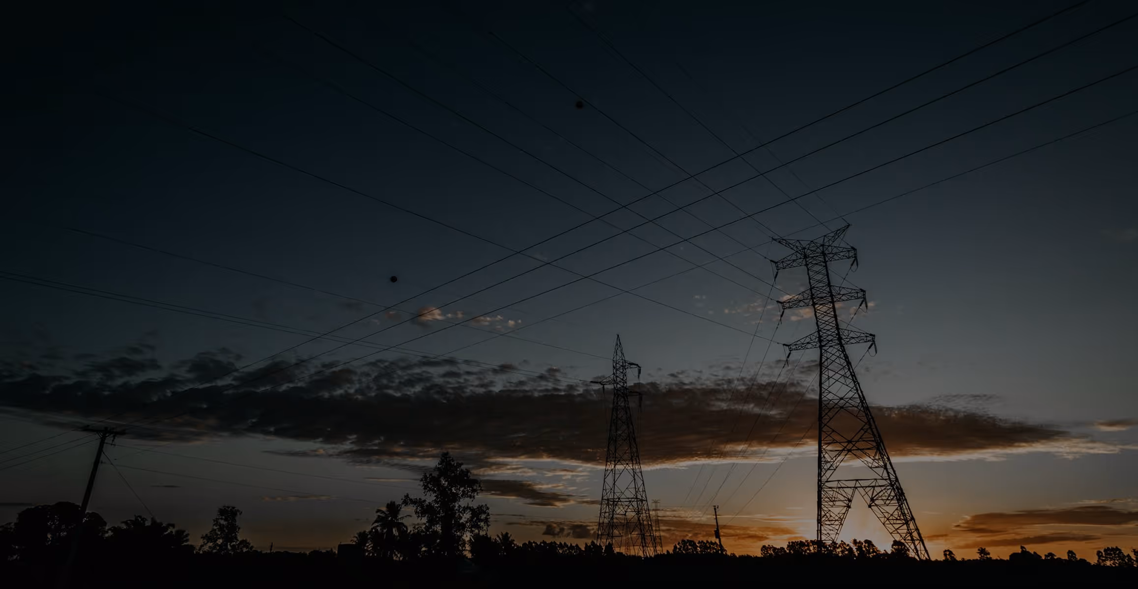 Silhouettes of electricity transmission towers and power lines against a sunset sky with scattered clouds.