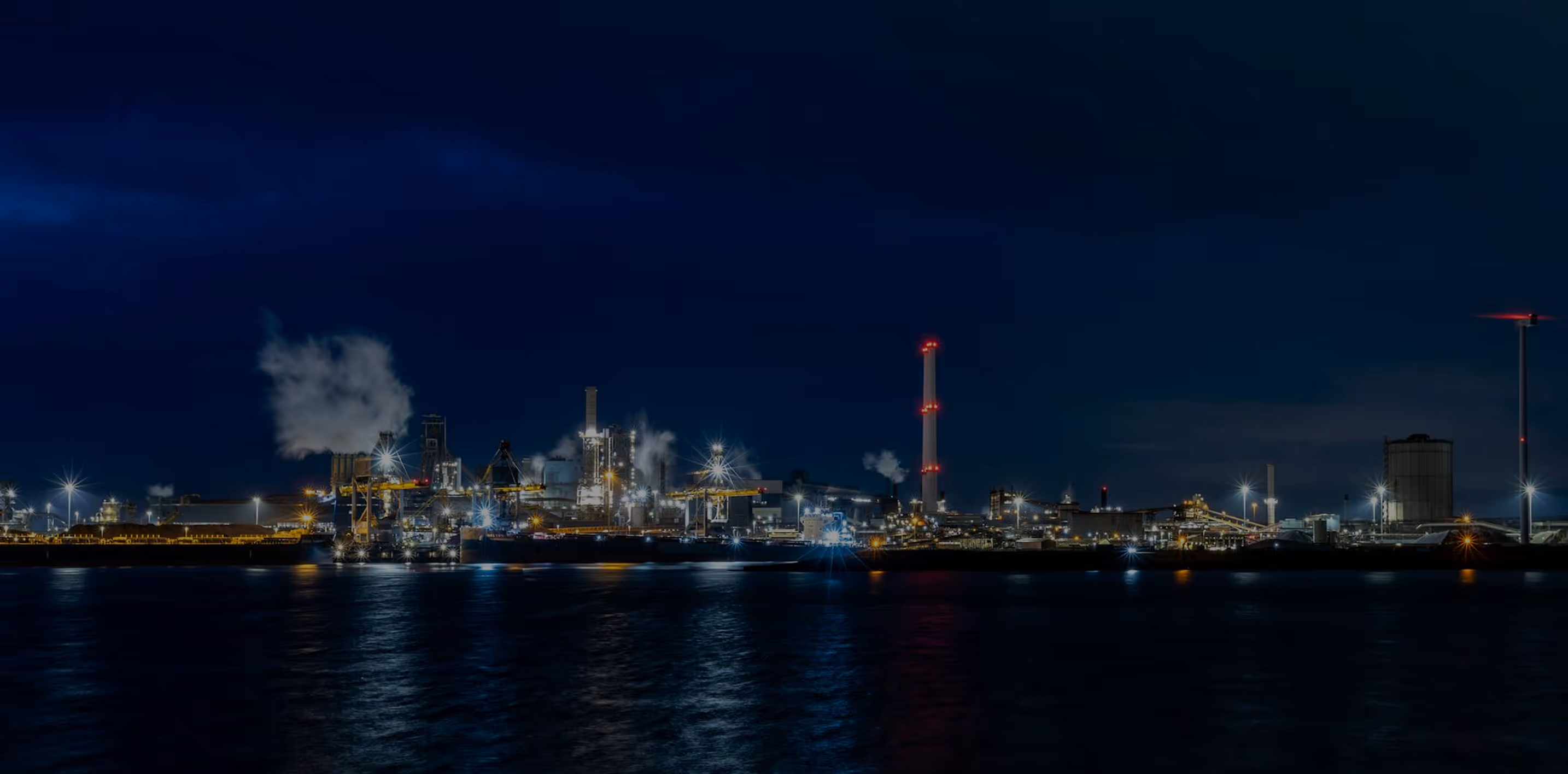 Industrial complex illuminated at night with lights reflecting on water and smoke rising from chimneys.