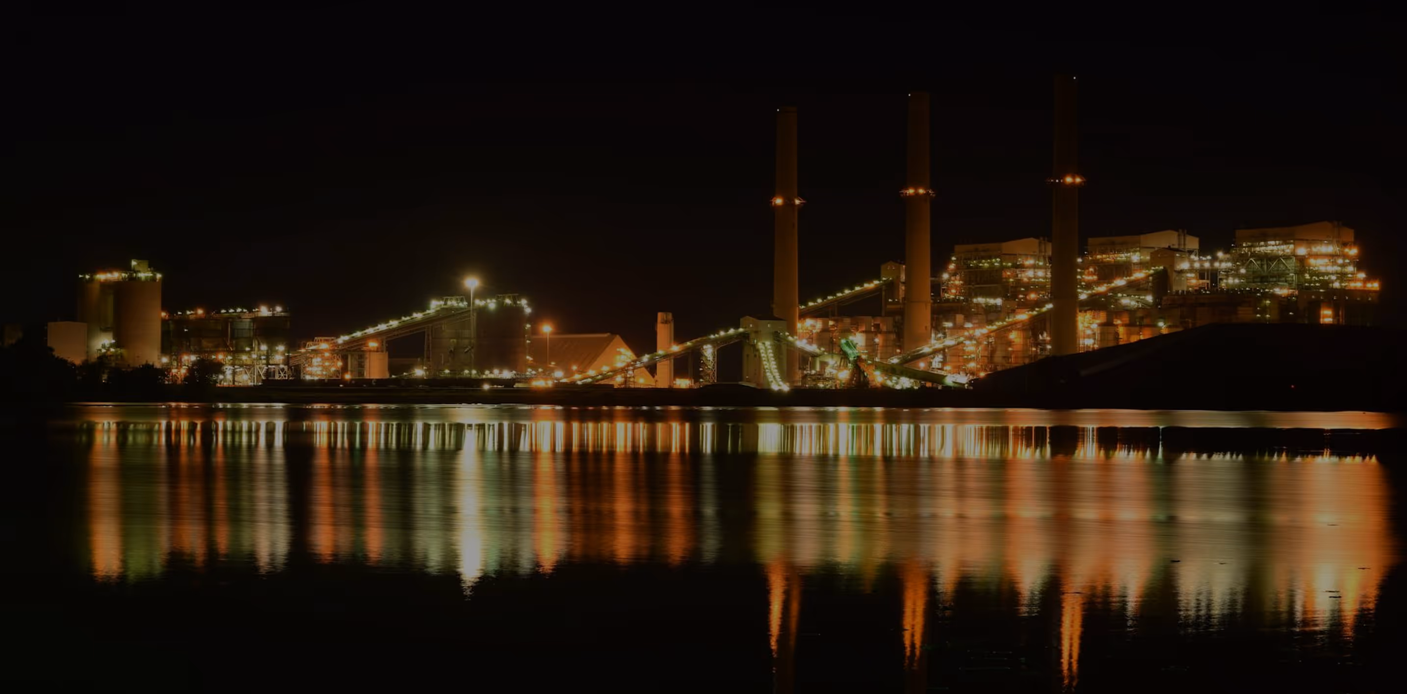 Illuminated industrial plant with tall chimneys reflecting on calm water at night.