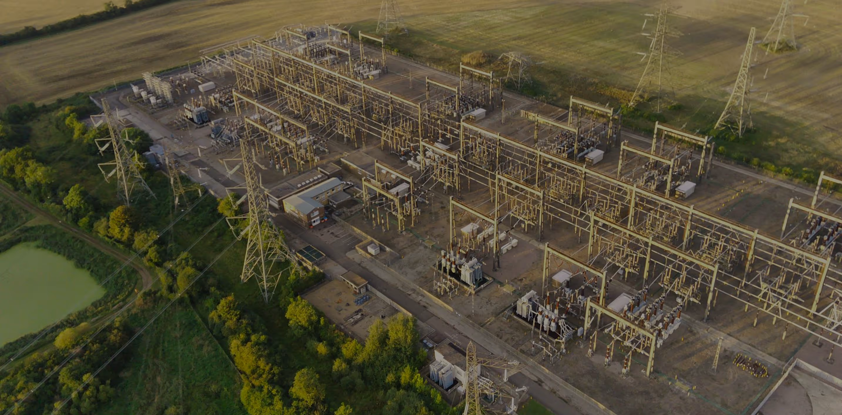 Aerial view of an electrical substation with transformers and power lines surrounded by green fields and trees.