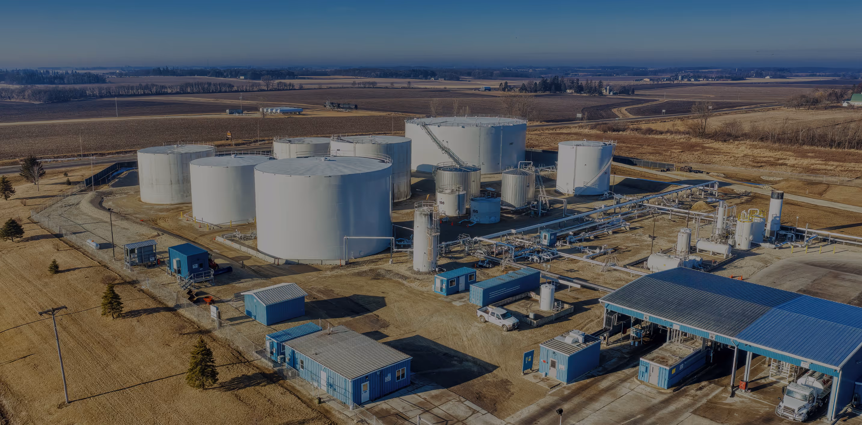 Aerial view of an industrial facility with large white storage tanks, blue utility buildings, and pipelines in a rural area.