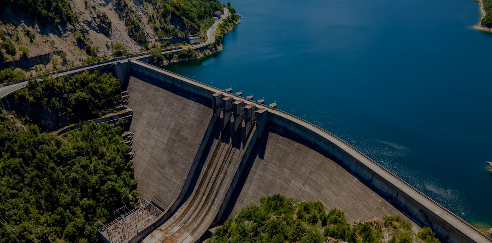A large concrete dam holding a reservoir surrounded by forested hills and rocky terrain.