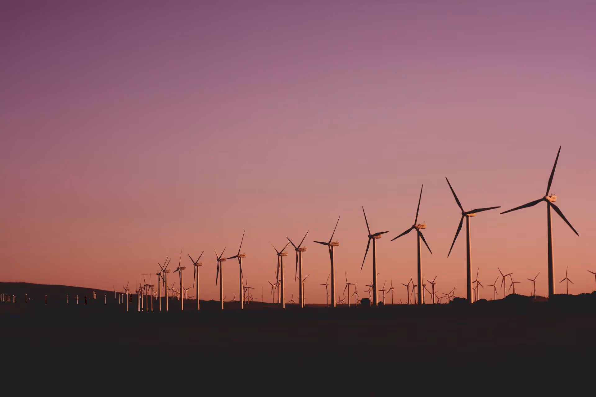 Line of wind turbines silhouetted against a purple and orange sunset sky over a dark landscape.
