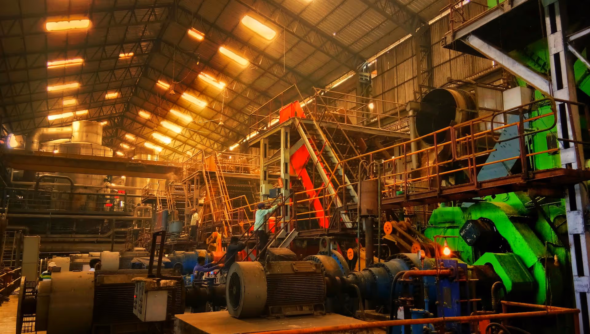 Industrial factory interior with large machinery, pipes, and workers operating equipment under bright ceiling lights.