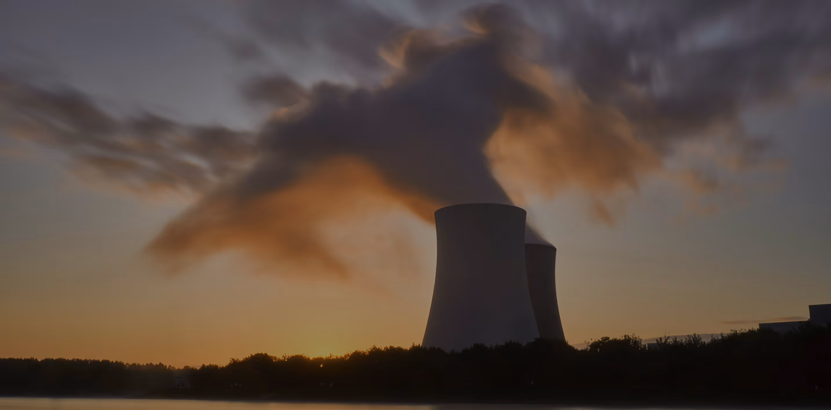 Cooling towers emitting steam against a sunset sky with trees and water in the foreground.