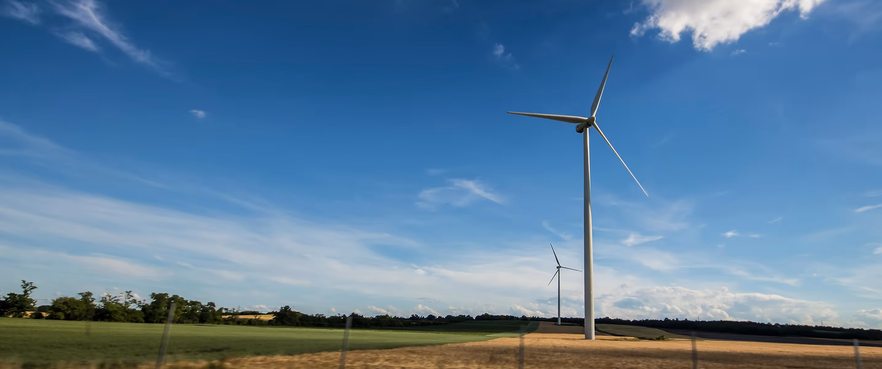 Two wind turbines in a rural field under a blue sky with scattered clouds.