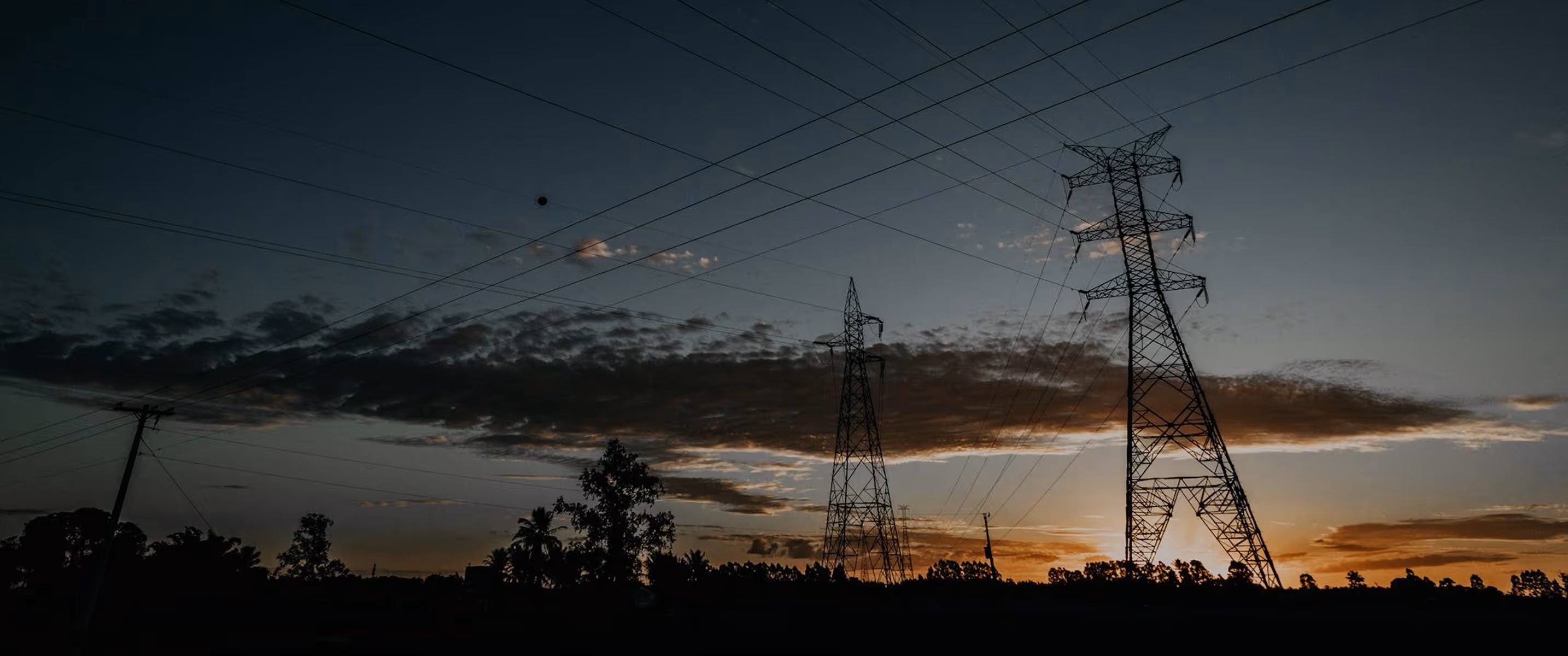 Silhouettes of electrical transmission towers and power lines against a sunset sky with scattered clouds.