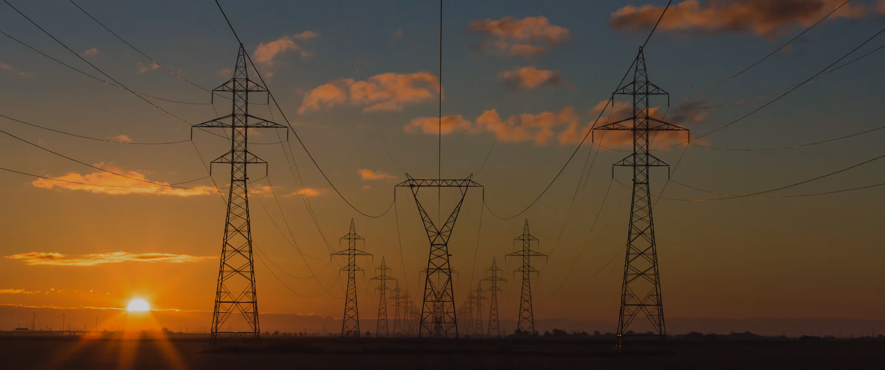 High-voltage power lines and transmission towers silhouetted against an orange sunset sky.