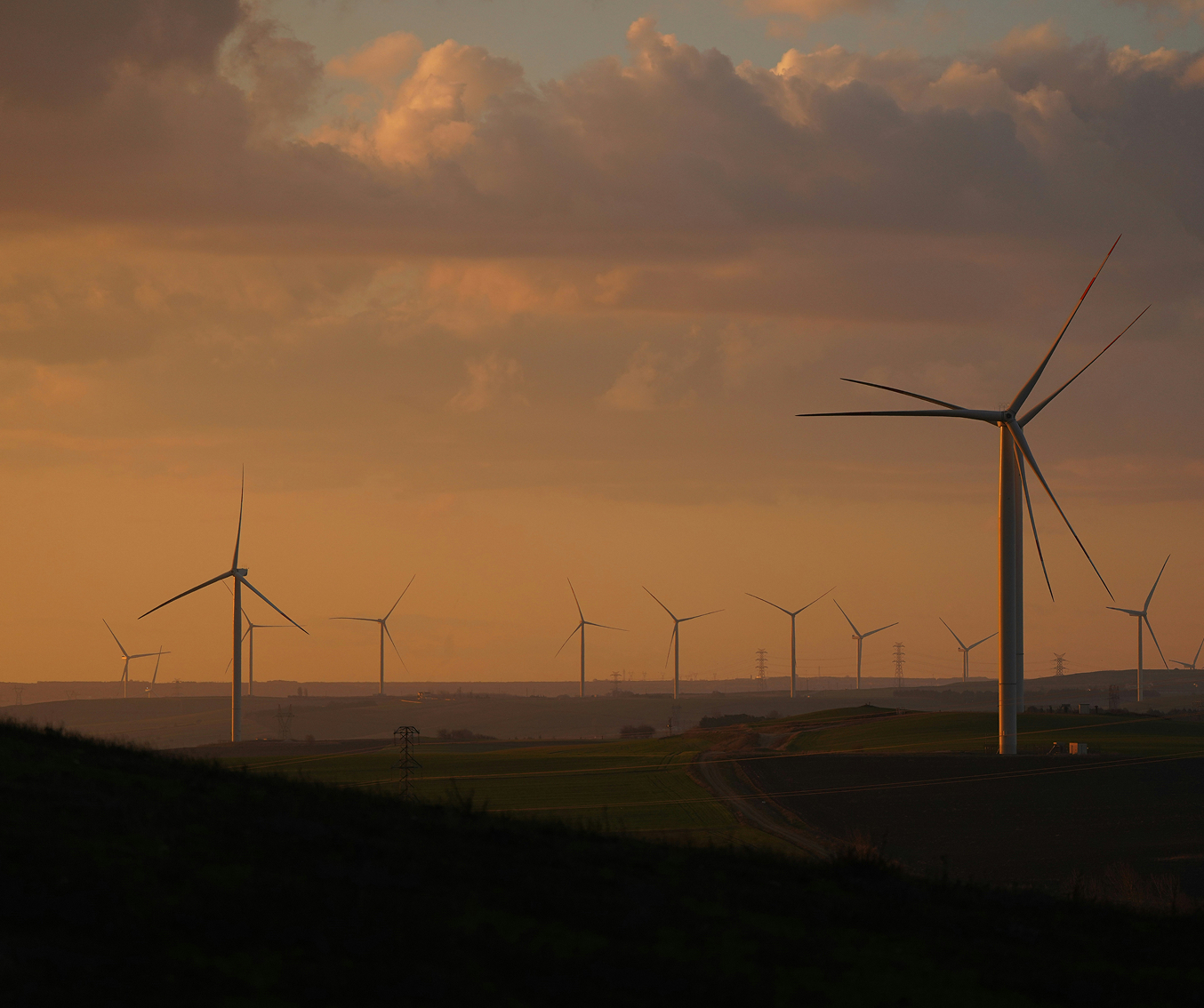 Wide landscape view of a wind farm with multiple wind turbines on a dry, hilly terrain under a cloudy sky.