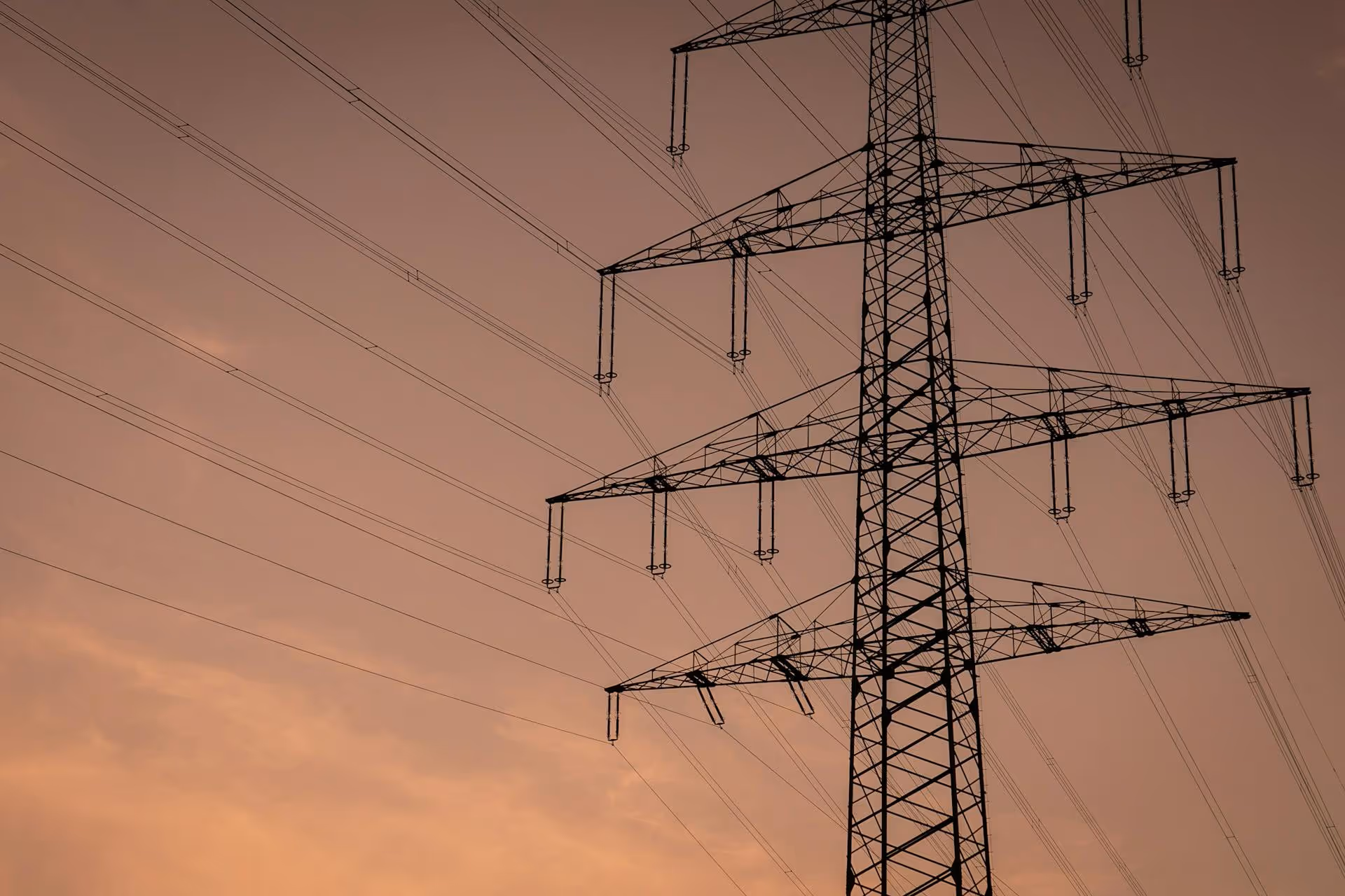 High voltage power transmission tower with electrical wires against an orange sky at dusk.