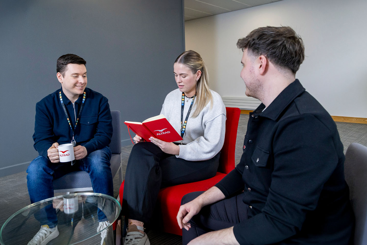 Three people seated indoors; one reads an Altrad-branded red book, another holds an Altrad mug.