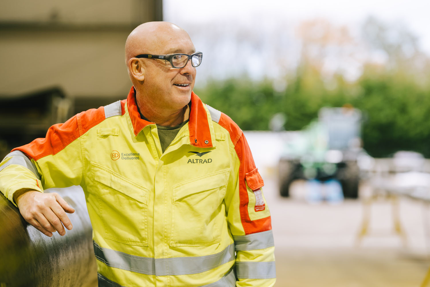 Smiling bald man wearing glasses and a high-visibility jacket with Altrad logo, outdoors.