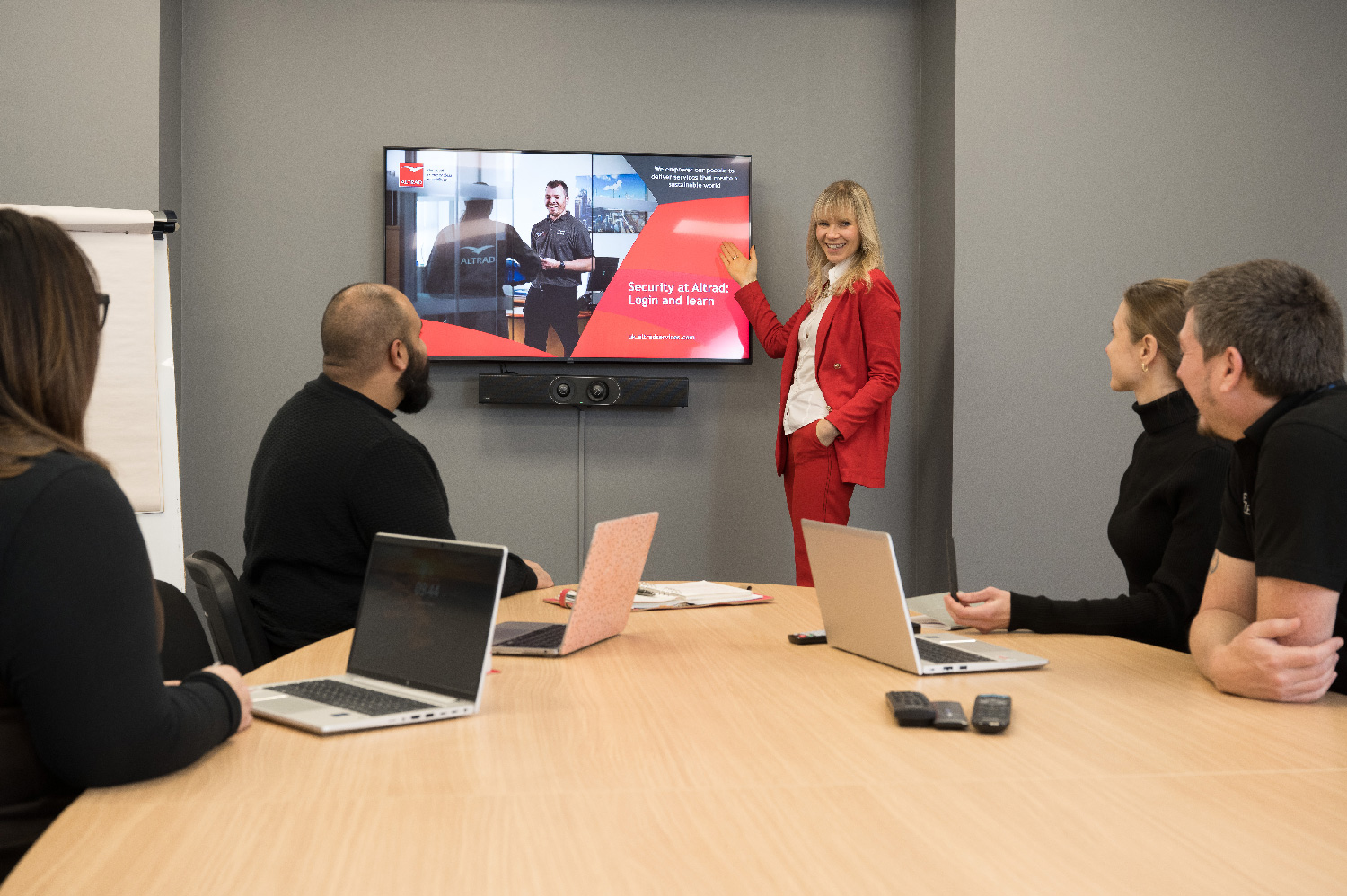 Woman in red suit presenting security training on screen to four people seated at a conference table with laptops.