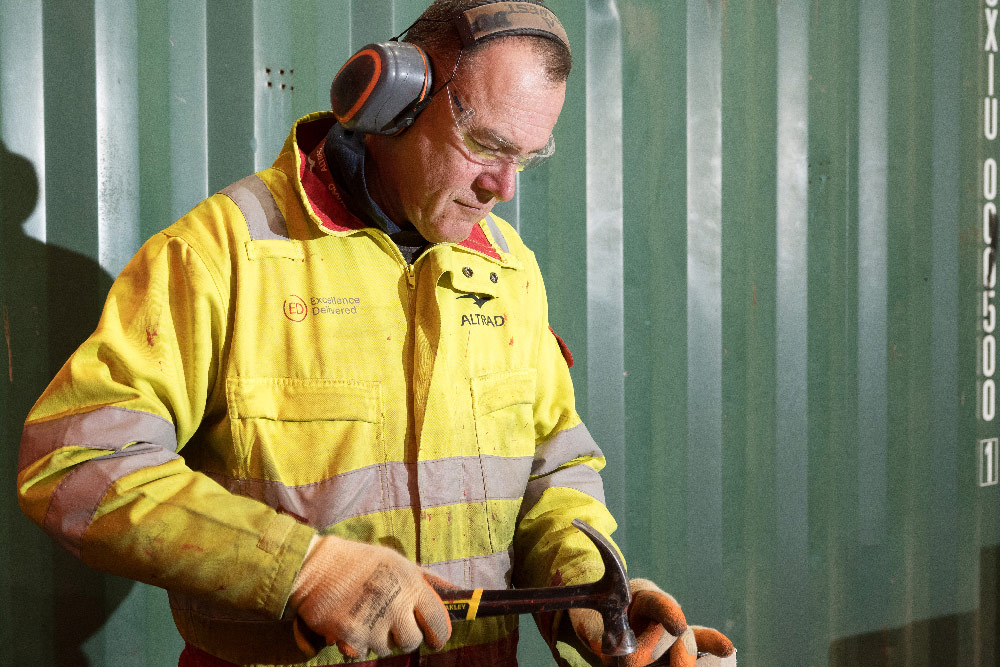 Man in yellow safety jacket and gloves using a hammer with ear protection and safety glasses.