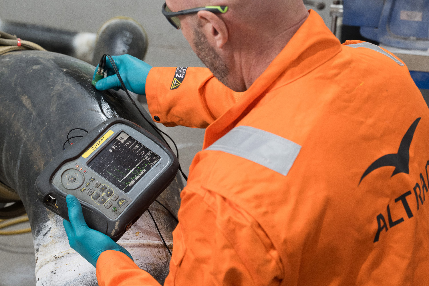 Technician in orange gear performing ultrasound inspection on a large pipe with a handheld device.