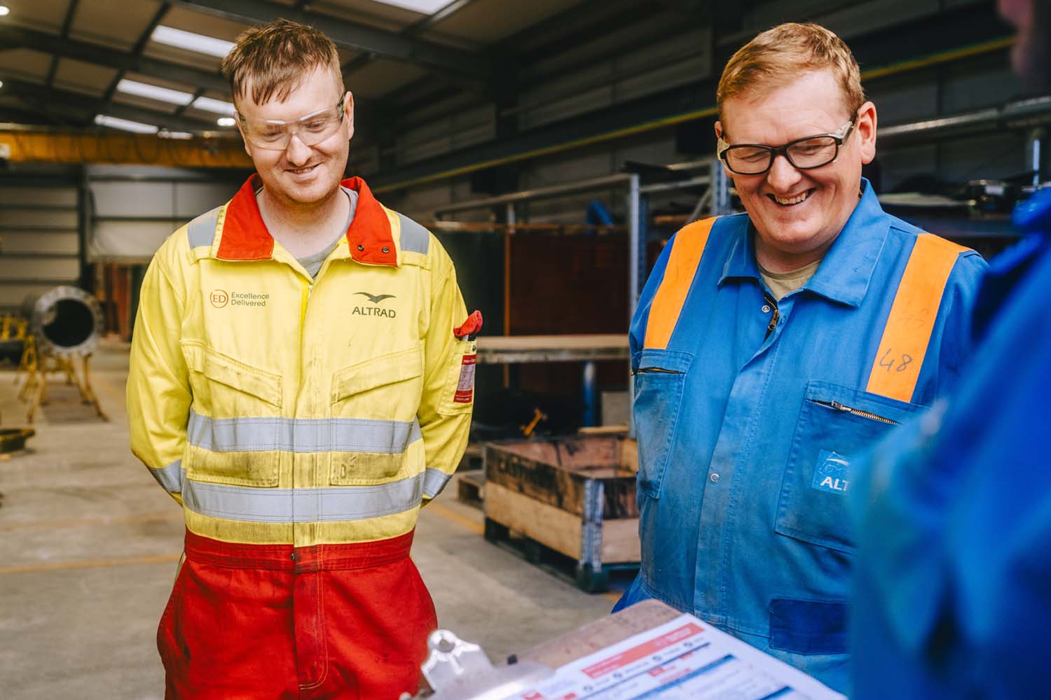 Two men in work coveralls smiling and looking at a clipboard in an industrial warehouse.