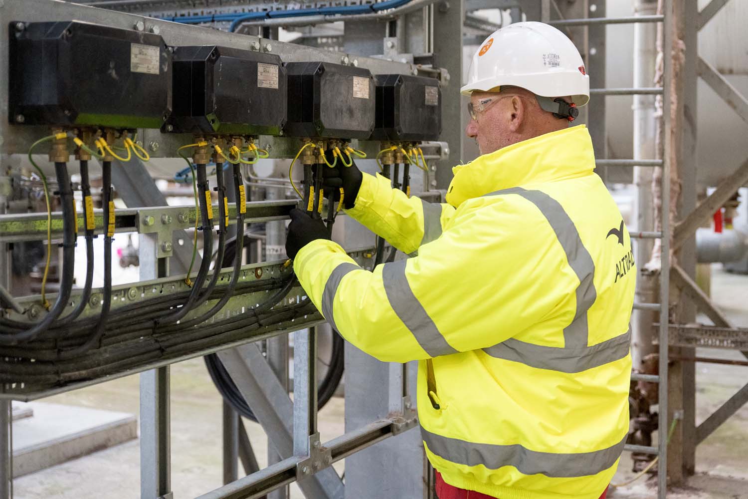 Worker in yellow safety jacket and white helmet adjusting industrial equipment with cables and connections.