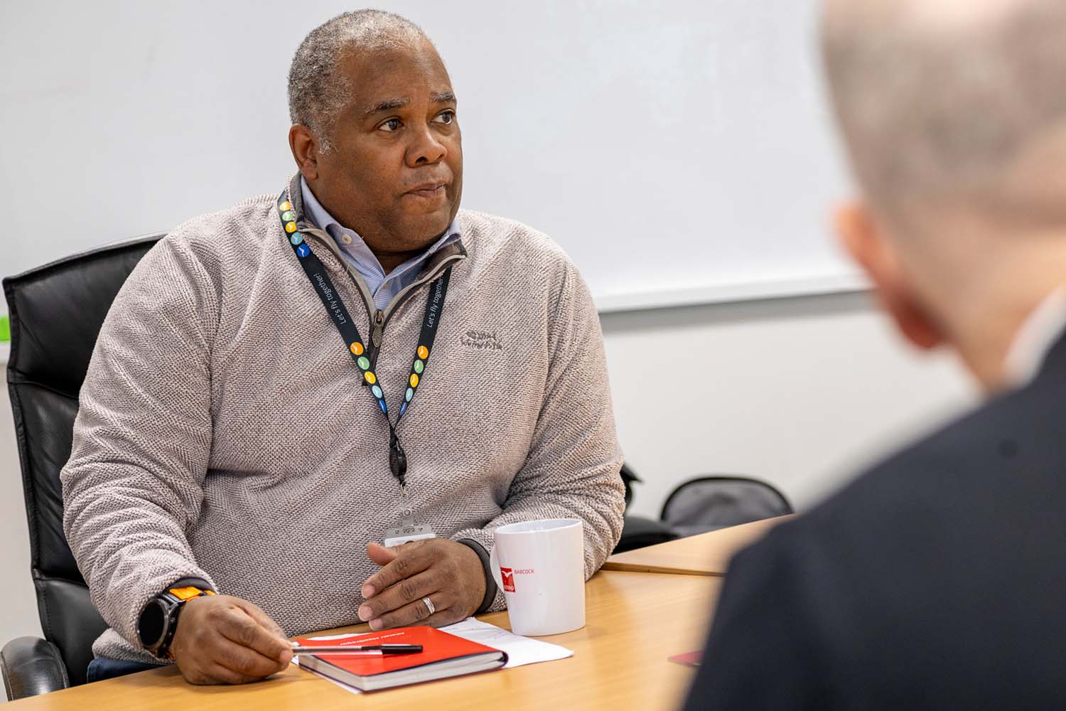 Man wearing a lanyard and sweater sitting at a table with a notebook, pen, and mug during a meeting.