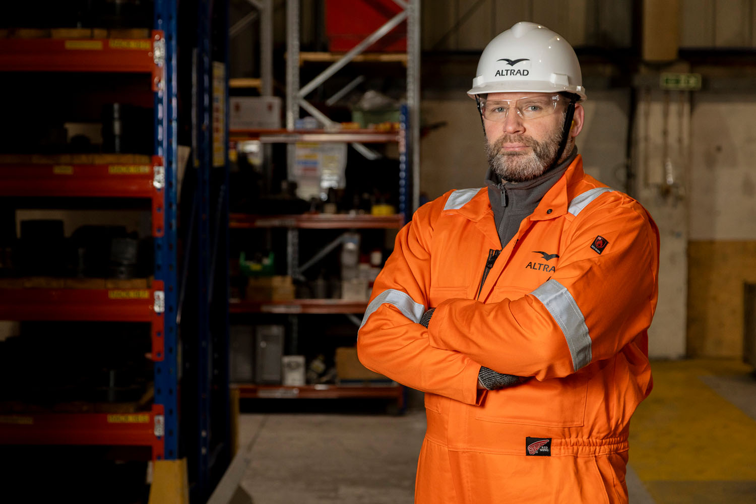 Man in orange safety coverall and white hard hat standing with arms crossed in warehouse.