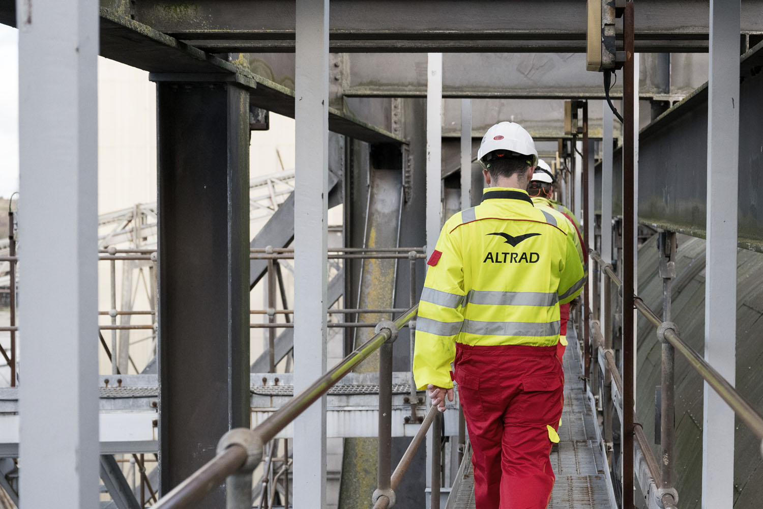 Workers wearing ALTRAD safety gear and helmets walking on an industrial metal walkway.