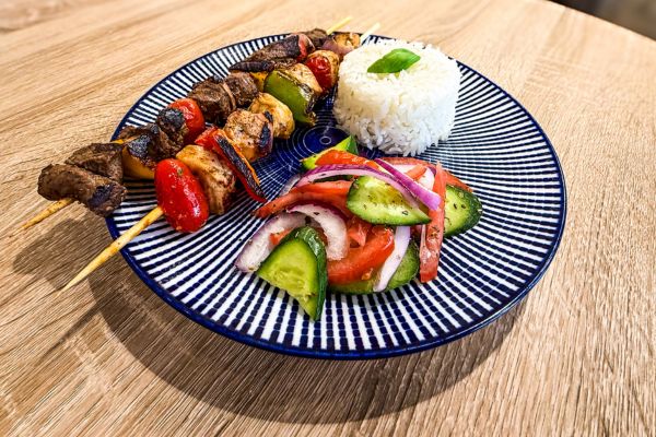 Plate with two grilled kebabs, a serving of white rice garnished with a basil leaf, and a fresh salad of cucumber, tomato, and red onion slices.