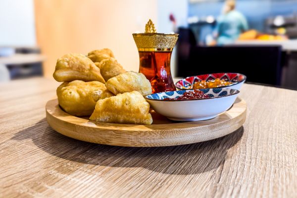 Round wooden tray with fried pastries, a patterned bowl of sauce, and a glass of tea on a wooden table.