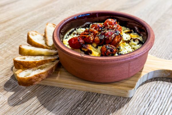Baked dish with roasted cherry tomatoes and herbs in a brown ceramic bowl served with slices of bread on a wooden board.