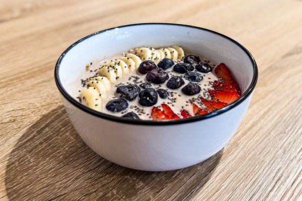 White bowl on wooden surface filled with creamy yogurt topped with sliced bananas, blueberries, strawberries, and chia seeds.