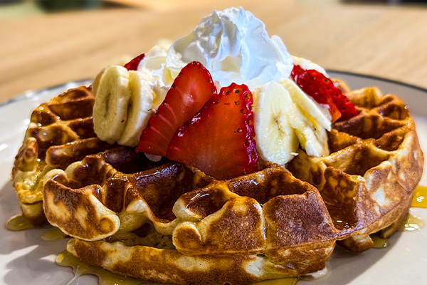 Golden-brown waffle topped with sliced strawberries, bananas, whipped cream, and drizzled with syrup on a white plate.