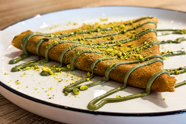 Triangular pistachio crepe on a white plate drizzled with green pistachio sauce and sprinkled with crushed pistachios.