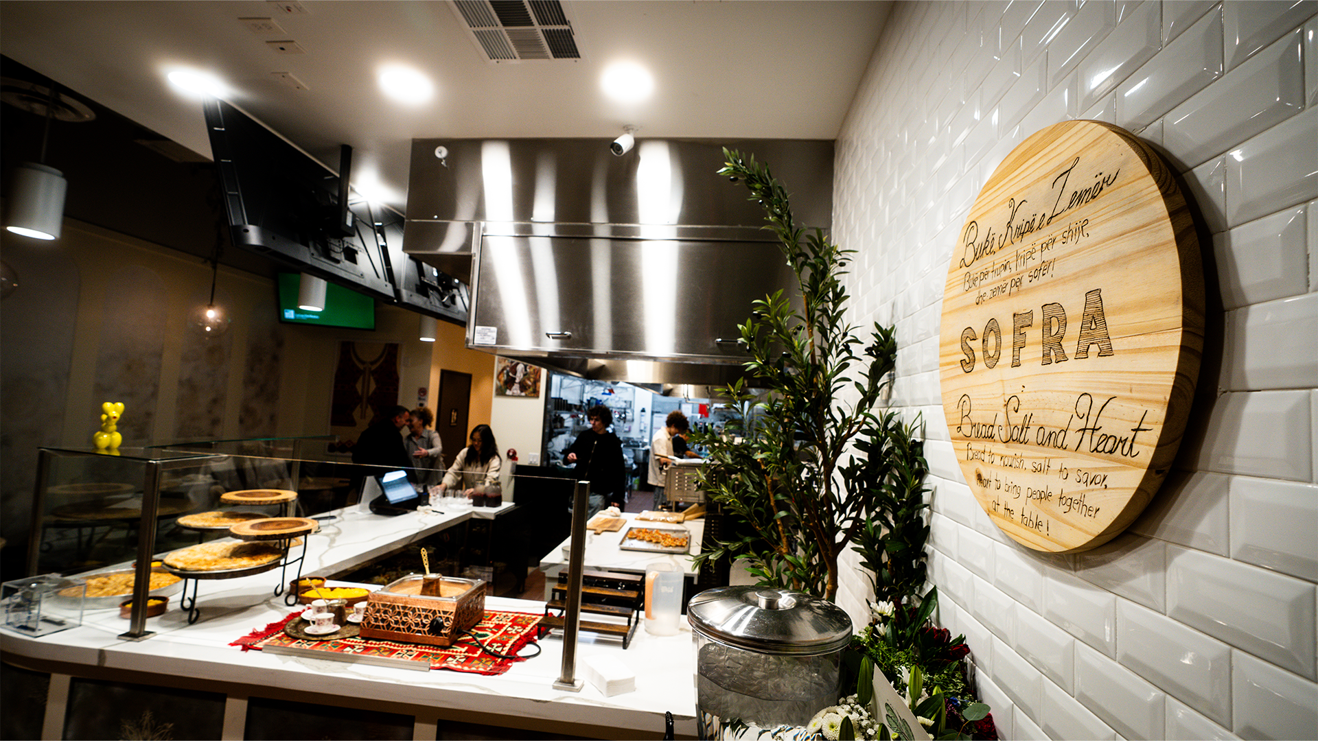 Interior of Sofra restaurant showing food service counter with pastries, a wooden sign with restaurant name on white tiled wall, and staff working in the kitchen in the background.