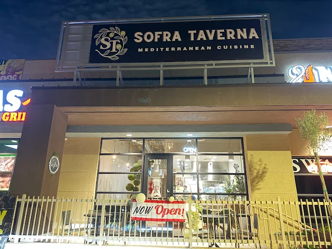 Nighttime view of Sofra Taverna Mediterranean Cuisine storefront with a 'Now Open!' banner and illuminated interior.